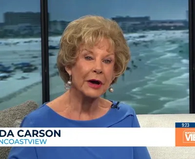 Linda Carson speaking on Suncoast View with a beach and ocean visible through windows behind her.