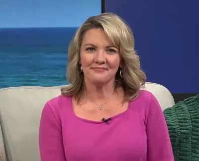 Woman with blonde hair wearing a pink top sitting on a light sofa with a green cushion and ocean background