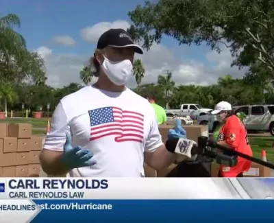 Man wearing American flag shirt and face mask speaking at an outdoor event with volunteers and boxes behind.