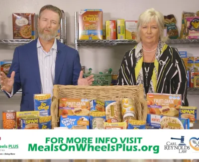 Two presenters stand behind a table of canned and boxed food with shelves of groceries in background promoting Meals on Wheels Plus.
