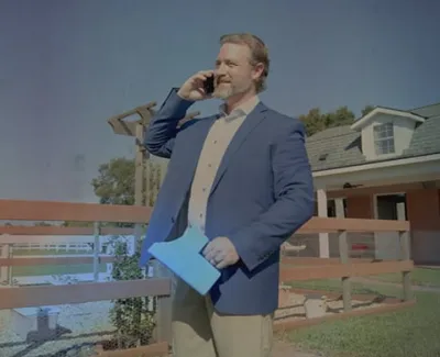 Businessman in blazer talking on phone while holding documents outside near a wooden fence and house.