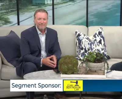 Two men in suits sitting on a couch during a televised talk show segment with beach scenery backdrop.