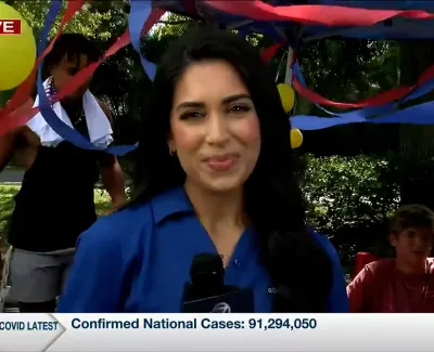 Female reporter in blue shirt stands in front of colorful streamers and balloons during a live outdoor broadcast.