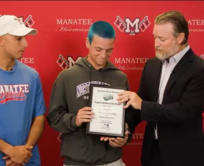 Student receiving Student Athlete of the Month award with two men in front of Manatee Hurricanes backdrop