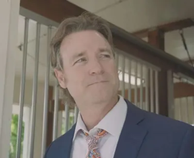 Man in a blue suit with a patterned tie looking to the side in a bright indoor setting with large windows.