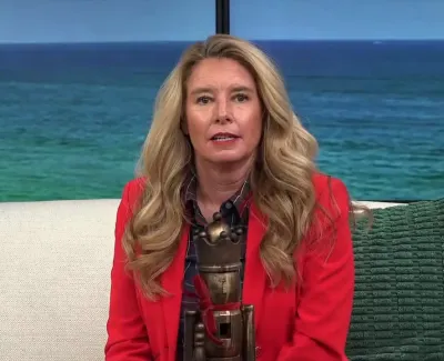 Woman in red blazer sitting on white couch with green pillow and ocean view behind her