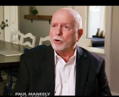 Elderly man in black suit and white shirt speaking in a well-lit dining room with wooden table and chairs.