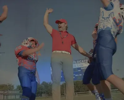 Youth football coach and players in blue and red uniforms celebrate on the field under a clear sky.