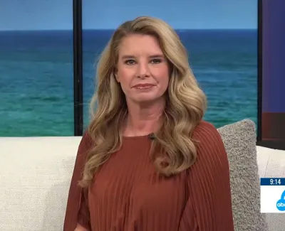 Woman with long blonde hair seated indoors with ocean view behind, wearing rust-colored blouse, news logo on screen