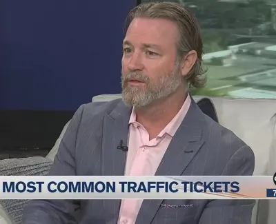 Man in a gray suit discussing common traffic tickets on a news show with a city view background.