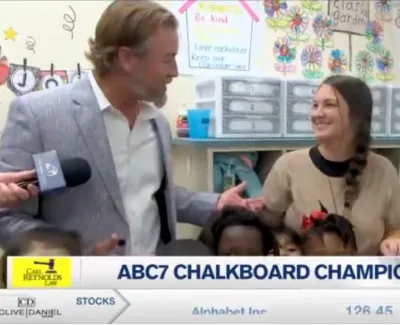 News reporter interviews a teacher surrounded by young students in a colorful classroom setting.