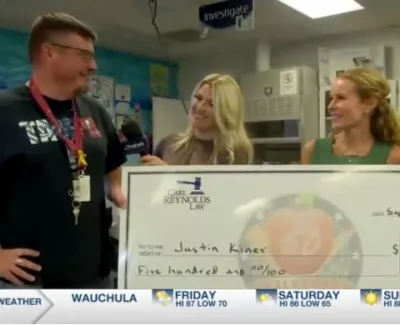 Man receiving oversized check from two smiling women during a local news segment in a classroom setting.