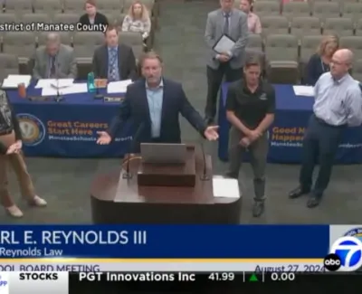 School board meeting with Carl E. Reynolds III speaking at a podium and others standing nearby in a conference room