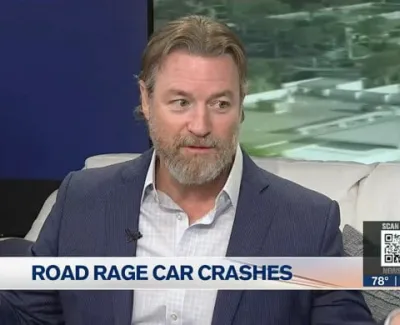 Man in suit discussing road rage car crashes on a news interview set with a cityscape background.