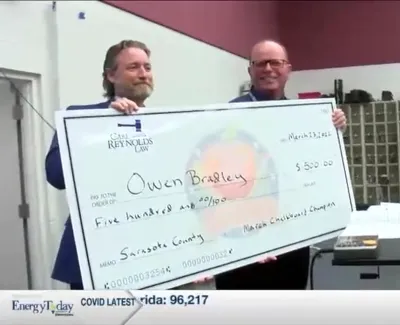 Two men holding a large check for Owen Bradley awarding $500 at a music event with students and instruments.