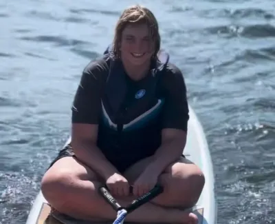 Smiling person in black wetsuit sitting cross-legged on paddleboard on calm water under sunlight.