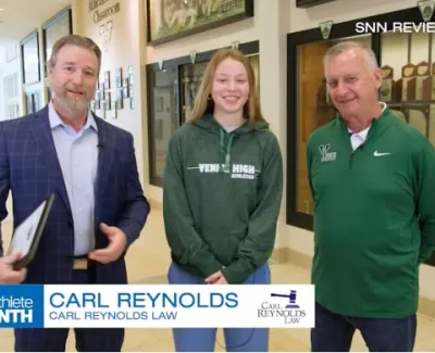 Student Athlete of the Month with Carl Reynolds and two individuals in a school hallway with trophies.