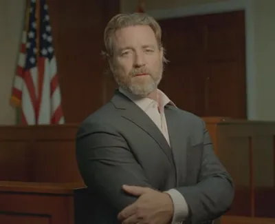 Man with beard in a suit standing confidently in a courtroom with an American flag in the background