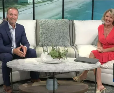 Two TV hosts sitting on white couches in a studio set with a coastal backdrop and marble coffee table.