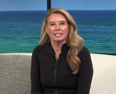 Woman with long blonde hair wearing black jacket sitting on white couch with ocean on screen behind her