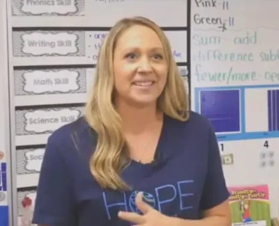 Female teacher in navy blue shirt stands in front of classroom whiteboard with educational charts and notes.