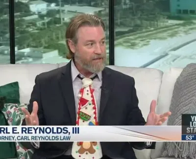 Attorney Carl E. Reynolds III speaking in a studio wearing a festive Christmas tie with holiday decorations in the background