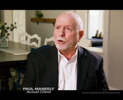 Elderly man in suit speaking in a bright dining room with a large wooden table and white chairs.