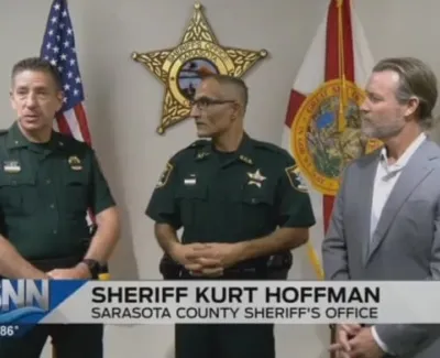 Sheriff Kurt Hoffman and officers speaking at Sarasota County Sheriff's Office with flags and badges in background
