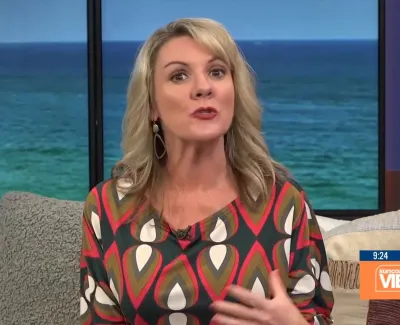 Woman with blonde hair wearing a colorful patterned top speaks on a TV show with ocean backdrop.