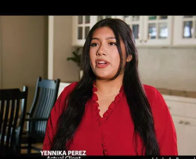 Woman with long dark hair wearing a red blouse speaking inside a bright dining room with black chairs and a wooden table.