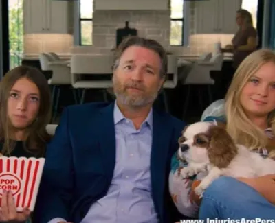 Man in suit sitting on couch with two girls, one holding popcorn, the other holding a small dog at home