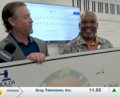Two men hold a large check in a classroom with music notes and charts in the background during a news segment.