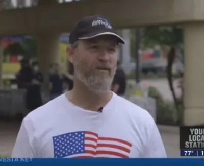 Man in a white shirt with American flag design and black cap being interviewed outdoors near a news station.