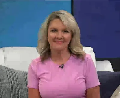 Smiling woman with blonde hair wearing a pink shirt sitting on a white couch with blue and gray pillows.