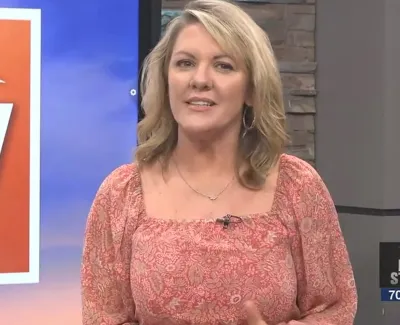 Female news anchor in coral patterned dress presents on local TV station with weather and time displayed