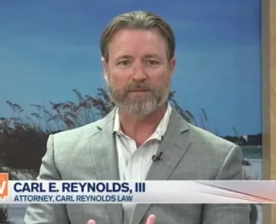 Attorney Carl E. Reynolds III in a gray suit speaking on Gulfcoast View television program set.