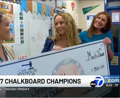 Three women smiling and holding a large $500 check in a colorful classroom setting with science posters.