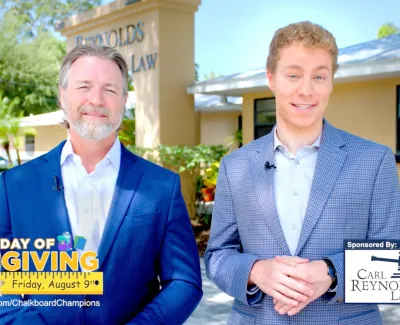 Two men in suits standing outdoors promoting Day of Giving event sponsored by Carl Reynolds Law on August 9th.