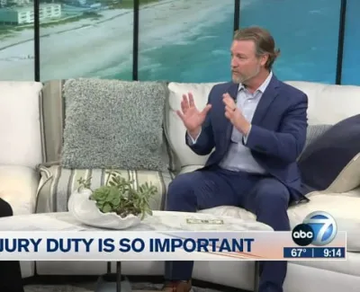 Two people discussing jury duty on a TV talk show set with beach view windows behind them.