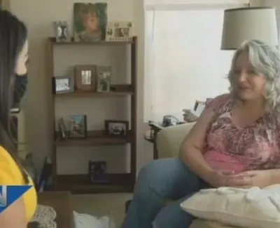 Two women in conversation in a living room, one interviewer wearing a yellow dress and face mask, the other seated on a couch.