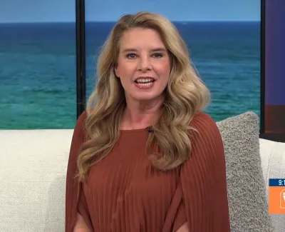 Blonde woman with wavy hair in a rust-colored blouse sitting on a couch with ocean background on TV show set.