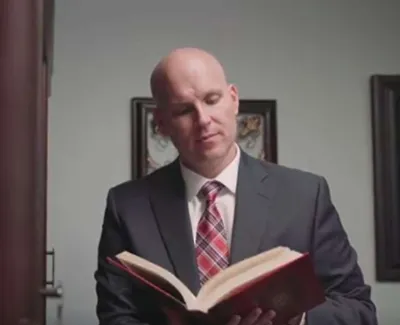 Bald man in a suit and plaid tie reading a large red book indoors with framed pictures on the wall.