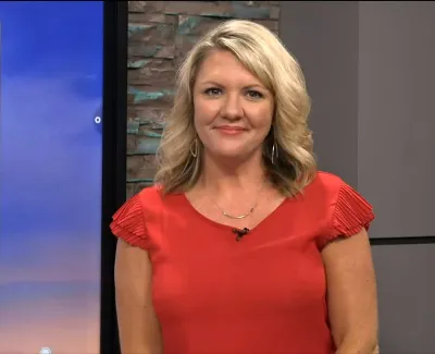 Smiling woman with blonde hair in a red blouse standing indoors in front of a monitor and stone wall.