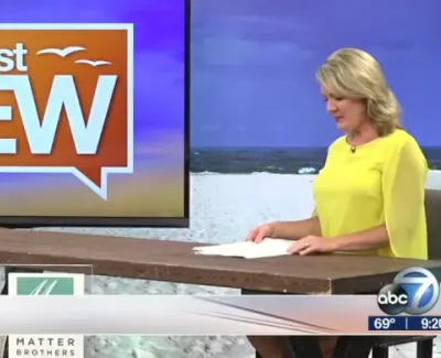 Suncoast View TV set with female host in yellow top preparing notes by a wooden table against a beach backdrop