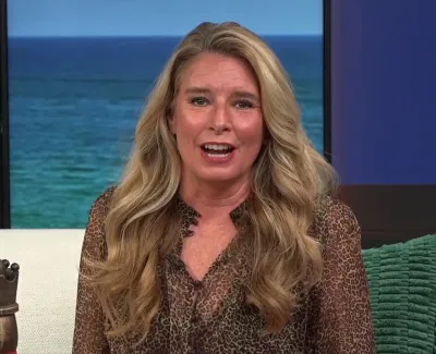 Blonde woman with long hair wearing a leopard print blouse sitting on a sofa near a large window showing the sea.