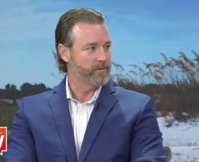 Man with beard and slicked-back hair wearing a blue suit and white shirt sitting in front of a beach backdrop with Suncoast View logo.