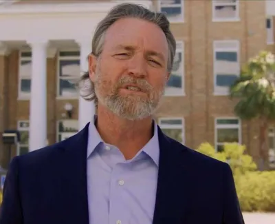 Middle-aged man with gray beard and blue blazer speaking in front of a brick institutional building with columns.
