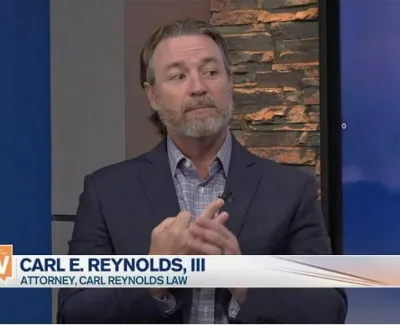 Attorney Carl E. Reynolds III speaking on Suncoast View TV show in a suit with brick background.