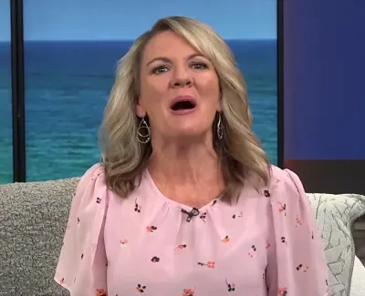 Blonde woman in pink floral blouse speaking on a couch with ocean view background and stone wall