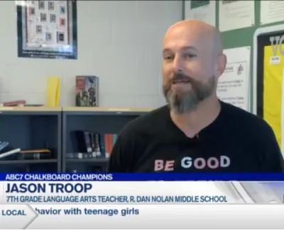 7th grade language arts teacher Jason Troop speaking in a classroom with bookshelves and vocabulary chart